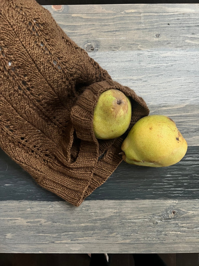 A rustic still life with ripe green pears on a textured wooden bowl and a knitted brown market bag, all set on a weathered wooden surface alongside unique jewelry.