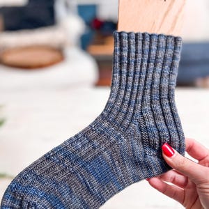 A hand with red nail polish holds a wooden sock blocker displaying a ribbed, blue and gray knitted sock—an ideal handmade gift. The softly blurred background reveals hints of furniture and home decor.