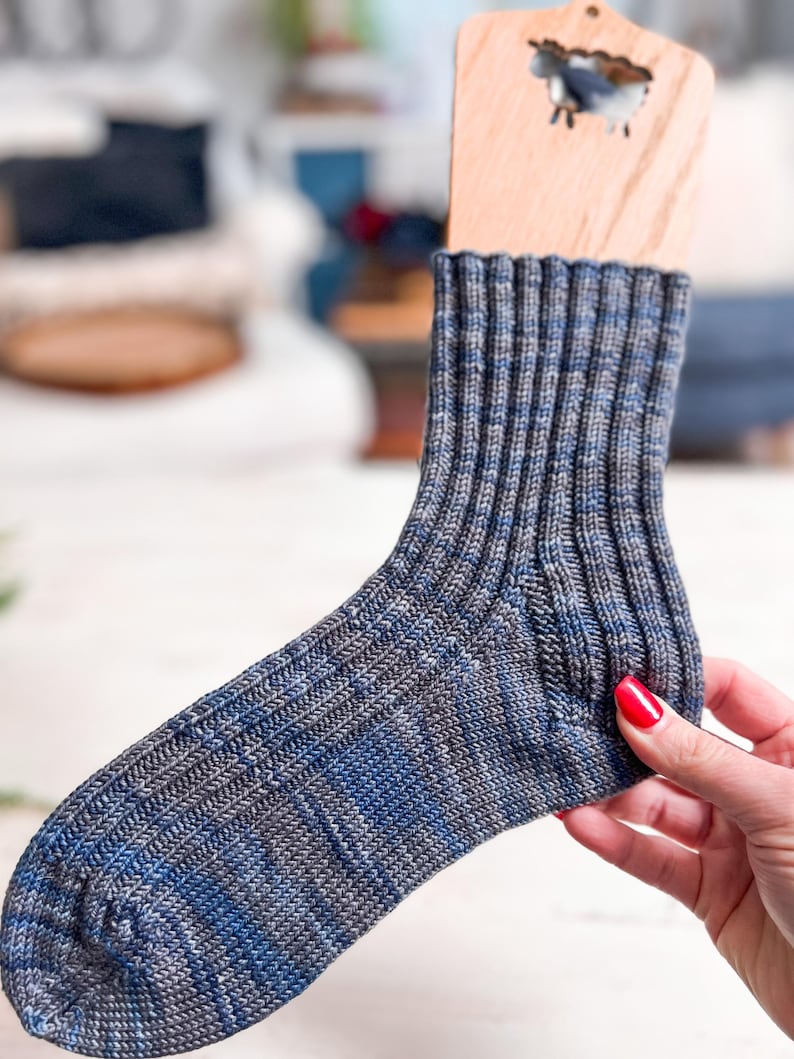 A hand with red nail polish holds a wooden sock blocker displaying a ribbed, blue and gray knitted sock—an ideal handmade gift. The softly blurred background reveals hints of furniture and home decor.