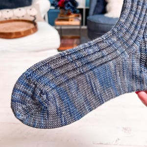 A close-up of a knitted sock in shades of blue and gray, showing fine ribbed texture, is held up in a cozy room with a white couch and blue chair—a perfect handmade gift for someone special.