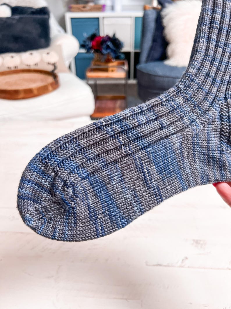 A close-up of a knitted sock in shades of blue and gray, showing fine ribbed texture, is held up in a cozy room with a white couch and blue chair—a perfect handmade gift for someone special.