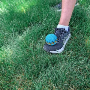 Blue crochet hacky sack resting on the toe of a sneaker on grass, ready to be kicked.