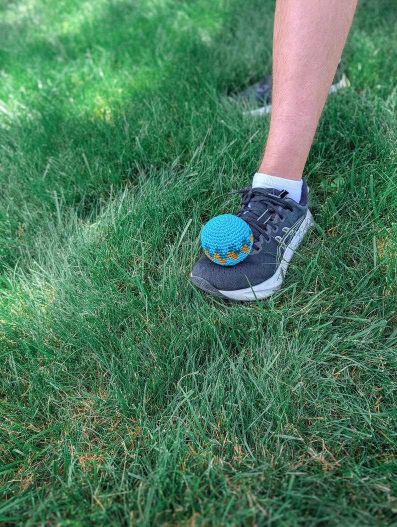 Blue crochet hacky sack resting on the toe of a sneaker on grass, ready to be kicked.