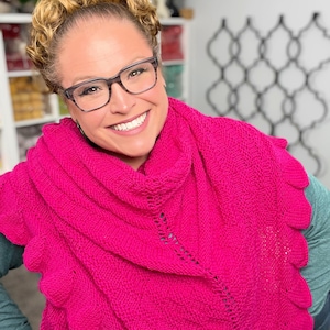 May include: A vibrant fuchsia knitted shawl with a scalloped edge. The shawl is wrapped around the shoulders. The person in the image is wearing glasses and a green long-sleeve shirt.