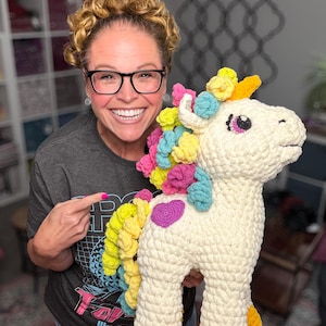 May include: A large, cream-colored crocheted unicorn plush toy featuring a vibrant, multi-colored mane and tail in shades of pink, yellow, blue, and green. The unicorn has a purple heart-shaped patch on its side and bright yellow hooves.