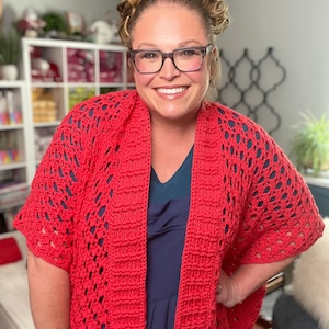May include: A vibrant red, open-weave crochet cardigan. The short-sleeved garment features a textured pattern and drapes over a dark blue top. The cardigan is a bold statement piece, perfect for layering.