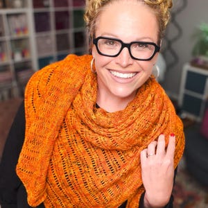 May include: An orange knitted shawl with a textured, openwork design. The shawl is draped around the shoulders. The color is a vibrant mix of orange and yellow. The shawl appears to be handmade.