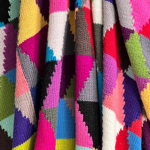 Peut inclure: Une couverture en tricot colorée avec un motif patchwork présentant différentes nuances de rose, bleu, vert, jaune, rouge, violet, blanc, noir et gris. La couverture est faite de fil doux et texturé et présente un motif géométrique.