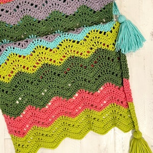A vividly colored Calor Crochet Wrap by Marly Bird laying flat on a rustic wood table, showcasing a wave crochet pattern and bright tassels.