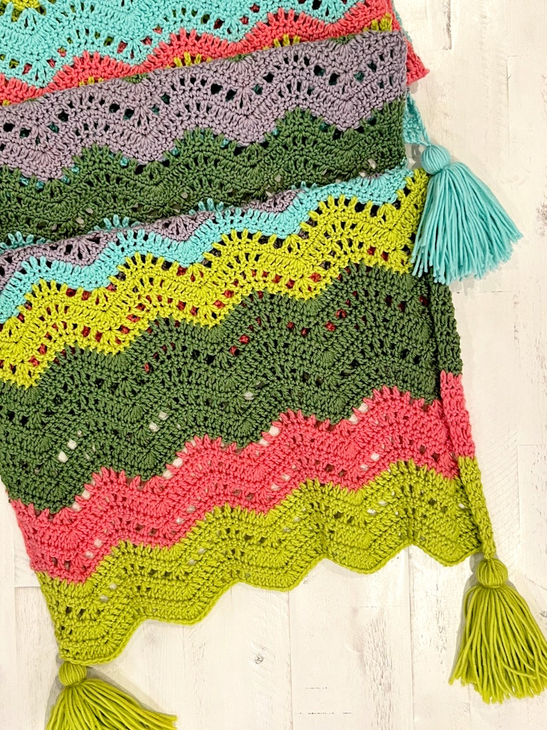 A vividly colored Calor Crochet Wrap by Marly Bird laying flat on a rustic wood table, showcasing a wave crochet pattern and bright tassels.