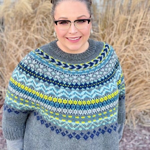May include: A woman wearing a gray knitted sweater with a blue, green, yellow, and white geometric pattern. She is wearing glasses and has her hair pulled back.