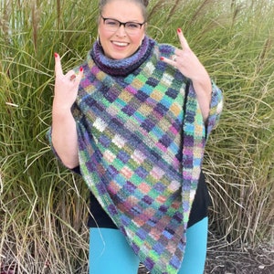 Marly Bird wearing the Check Me Out Gingham Plaid Crochet Poncho. The poncho is a rich tapestry of multicolored squares in hues of purple, green, and blue, with subtle tweed textures. The cowl neckline of the poncho showcases the gingham plaid stitch