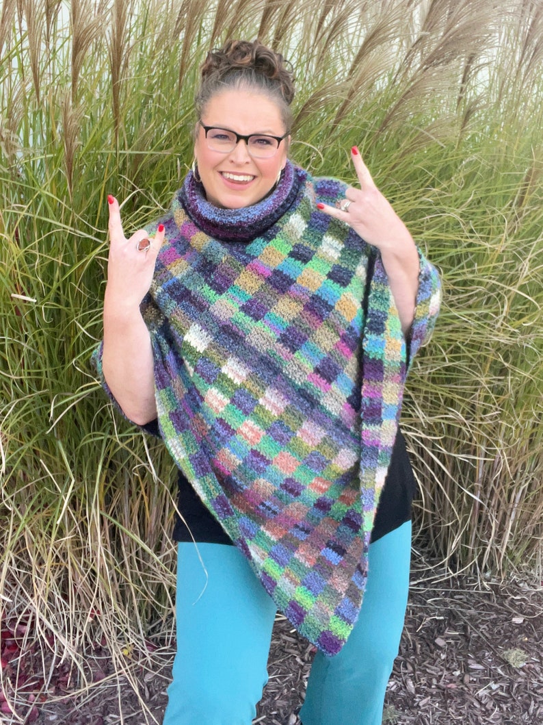 Marly Bird wearing the Check Me Out Gingham Plaid Crochet Poncho. The poncho is a rich tapestry of multicolored squares in hues of purple, green, and blue, with subtle tweed textures. The cowl neckline of the poncho showcases the gingham plaid stitch