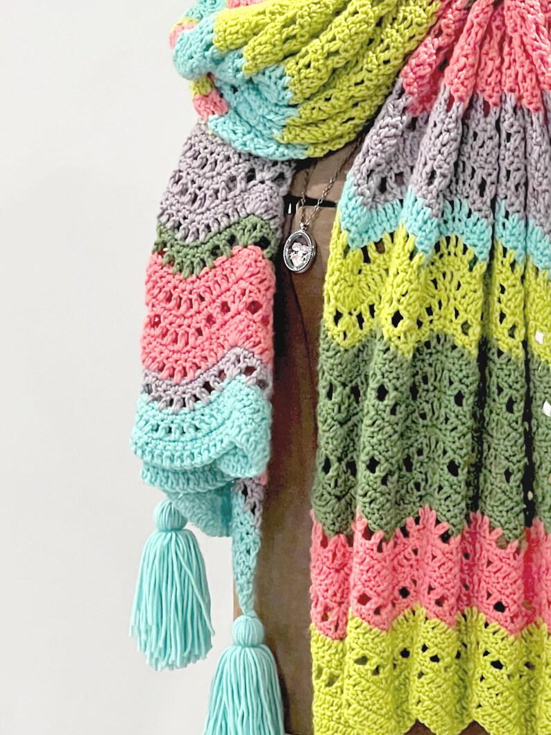 A vividly colored Calor Crochet Wrap by Marly Bird draped over a mannequin, showcasing a wave crochet pattern and bright tassels.