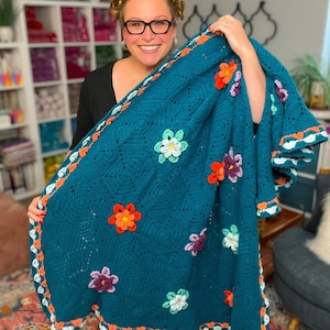 May include: A teal crochet blanket featuring a textured diamond pattern and scattered three-dimensional floral appliqués in orange, purple, and light green. The blanket is finished with a decorative border of orange and light green crochet motifs.