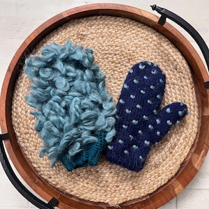 May include: Two handmade mittens displayed on a round wooden tray with a woven jute base. One mitten is a textured teal color, and the other is navy blue with light blue heart-shaped accents. The tray has a dark metal handle.