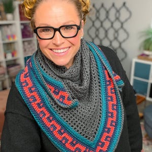 May include: A gray crochet shawl with a geometric pattern in teal and coral. The shawl is triangular and appears to be handmade. The design features a repeating pattern along the edges, creating a decorative border.