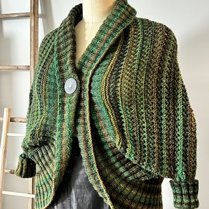 An elegantly draped, hand-knit cocoon cardigan displayed on a mannequin, showcasing rich hues of green with a subtle variegated pattern, chunky ribbed collar, and a large decorative button at the front. Marly Bird Know Your Worth Knit Cocoon Cardigan