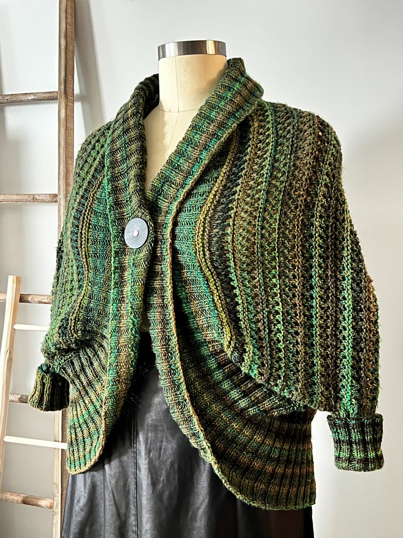 An elegantly draped, hand-knit cocoon cardigan displayed on a mannequin, showcasing rich hues of green with a subtle variegated pattern, chunky ribbed collar, and a large decorative button at the front. Marly Bird Know Your Worth Knit Cocoon Cardigan