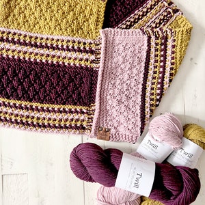 three color knit bandana cowl flat on a wood surface to show the 3d seam. This seam and the construction of the cowl form a point in front when wearing it. The cowl is made in light pink, wine purple, and a golden wheat color all shown in hank form.