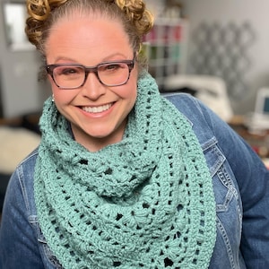 May include: A teal crochet shawl with a delicate openwork pattern. The shawl is worn by a person with curly hair and glasses.