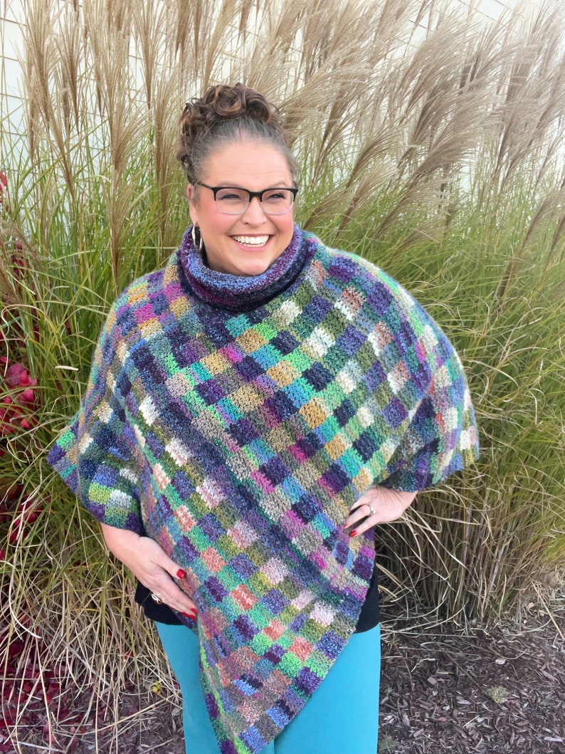 Marly Bird wearing the Check Me Out Gingham Plaid Crochet Poncho. The poncho is a rich tapestry of multicolored squares in hues of purple, green, and blue, with subtle tweed textures. The cowl neckline of the poncho showcases the gingham plaid stitch