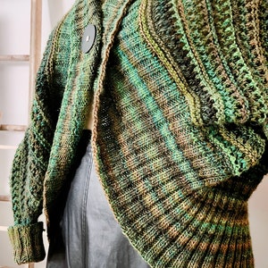 An elegantly draped, hand-knit cocoon cardigan displayed on a mannequin, showcasing rich hues of green with a subtle variegated pattern, chunky ribbed collar, and a large decorative button at the front. Marly Bird Know Your Worth Knit Cocoon Cardigan