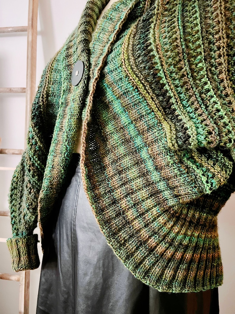 An elegantly draped, hand-knit cocoon cardigan displayed on a mannequin, showcasing rich hues of green with a subtle variegated pattern, chunky ribbed collar, and a large decorative button at the front. Marly Bird Know Your Worth Knit Cocoon Cardigan