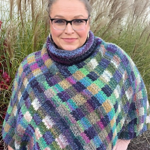 Marly Bird wearing the Check Me Out Gingham Plaid Crochet Poncho. The poncho is a rich tapestry of multicolored squares in hues of purple, green, and blue, with subtle tweed textures. The cowl neckline of the poncho showcases the gingham plaid stitch