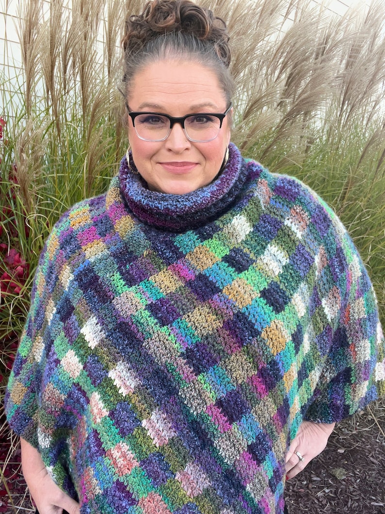 Marly Bird wearing the Check Me Out Gingham Plaid Crochet Poncho. The poncho is a rich tapestry of multicolored squares in hues of purple, green, and blue, with subtle tweed textures. The cowl neckline of the poncho showcases the gingham plaid stitch