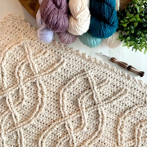 A crochet project in progress with a beautiful textured pattern, surrounded by skeins of colorful yarn and crochet tools, evoking a sense of creativity and craftsmanship for a unique handmade gift. Inishmore Crochet Cable Blanket - Marly Bird