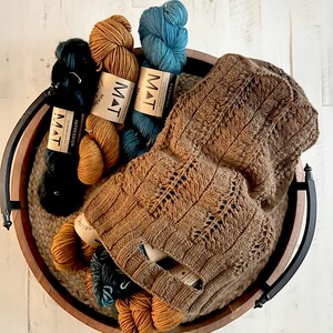 A rustic wooden basket filled with various colors of yarn and a knit market bag showcasing intricate patterns, placed on a white wooden surface, pattern available Marly Bird