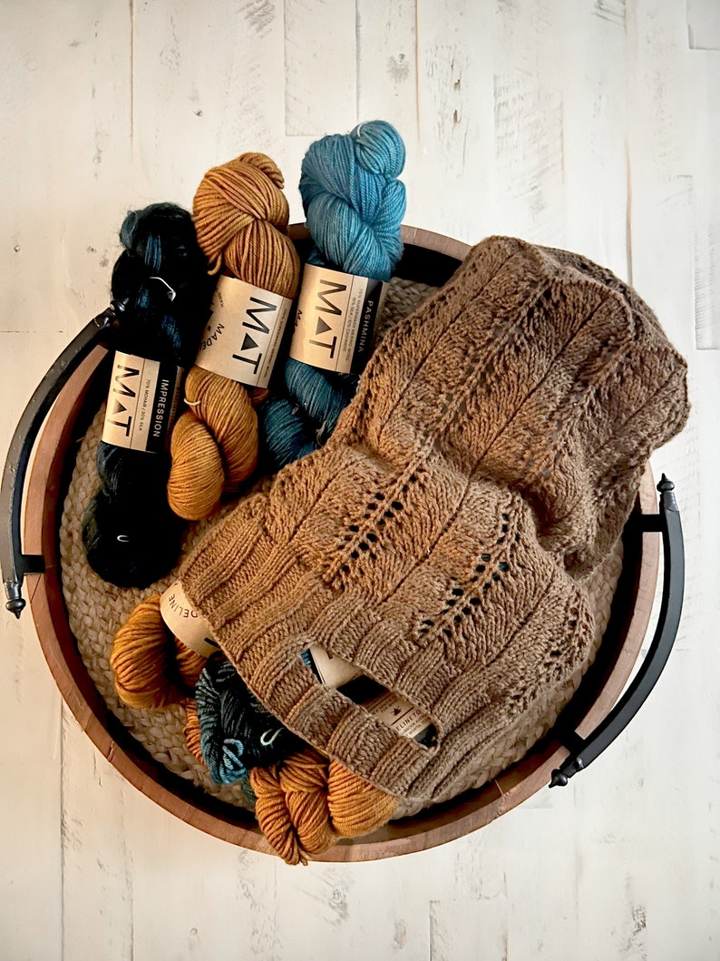 A rustic wooden basket filled with various colors of yarn and a knit market bag showcasing intricate patterns, placed on a white wooden surface, pattern available Marly Bird