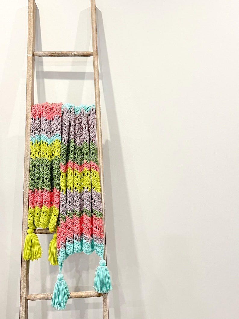 A vividly colored Calor Crochet Wrap by Marly Bird draped over a wood ladder that is leaning against a gray wall, showcasing a wave crochet pattern and bright tassels.