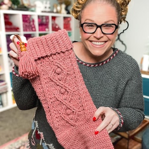 Puede incluir: Un calcetín navideño tejido de color rosa empolvado, con un diseño de punto trenzado y una borla. El calcetín tiene un puño doblado y un detalle de cuentas de madera en el lazo para colgar. Fondo: una habitación con estanterías blancas.