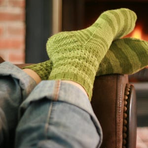 May include: A pair of green knitted socks with textured and striped patterns. The socks are worn with blue jeans, suggesting a cozy, relaxed setting. The background includes a brick wall and a blurred fireplace, creating a warm atmosphere.