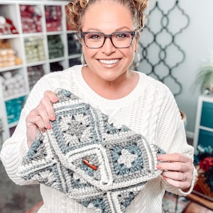 May include: A handmade crochet clutch in shades of gray, white, and blue. The clutch features a square pattern with a wooden button closure. The overall design is a mix of geometric shapes and soft textures, perfect for carrying essentials.