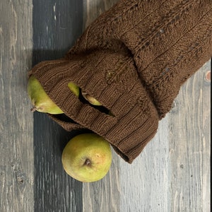 A rustic still life with ripe green pears on a textured wooden bowl and a knitted brown market bag, all set on a weathered wooden surface alongside unique jewelry.