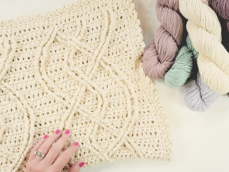 A crochet project in progress with a beautiful textured pattern, surrounded by skeins of colorful yarn and crochet tools, evoking a sense of creativity and craftsmanship for a unique handmade gift. Inishmore Crochet Cable Blanket - Marly Bird