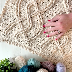 A crochet project in progress with a beautiful textured pattern, surrounded by skeins of colorful yarn and crochet tools, evoking a sense of creativity and craftsmanship for a unique handmade gift. Inishmore Crochet Cable Blanket - Marly Bird