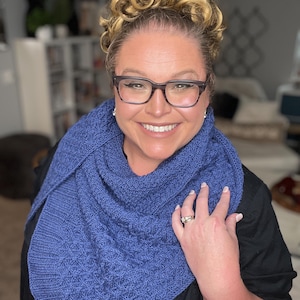May include: A woman wearing a blue knitted shawl with a cable knit pattern. She is wearing a black shirt and has her blonde hair styled in a bun with curls.