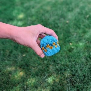 Hand holding a small blue crochet hacky sack with orange zigzag stripes above green grass.