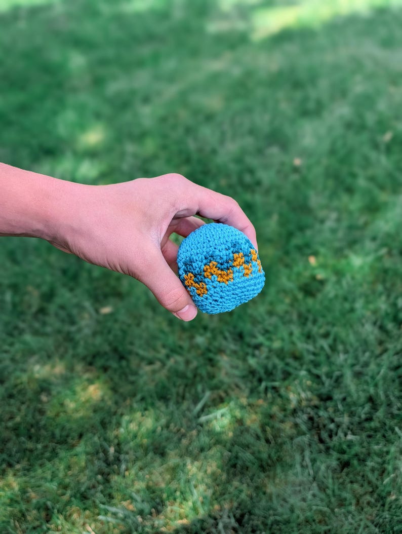 Hand holding a small blue crochet hacky sack with orange zigzag stripes above green grass.