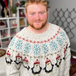 May include: A speckled, cream-colored sweater with a round neckline. The sweater features a pattern of blue snowflakes, red and pink accents, and black and white penguins wearing red hats. The sweater is made of a thick, warm material.