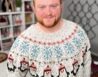 May include: A cream-coloured, speckled sweater with a round neckline. The sweater features a pattern of blue snowflakes, red and pink accents, and black and white penguins wearing red hats. The sweater is made of a thick, warm material.