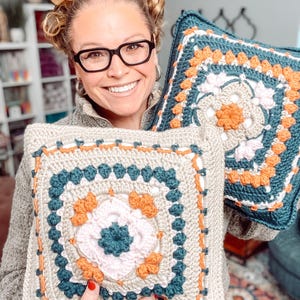 May include: Two square crochet pillows with floral designs. One pillow is held up, displaying a beige background with teal, orange, and pink floral patterns. The other pillow has a teal background with orange and pink floral patterns.