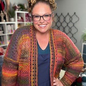 May include: A colorful, open-front crochet cardigan in shades of orange, red, yellow, and purple. The cardigan has a textured, open weave design. The person in the image is wearing the cardigan over a dark blue dress.