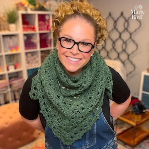 May include: A woman wearing a green crocheted shawl. The shawl is a triangular shape with an open, lacy design. The woman is smiling and wearing black-framed glasses. The background is blurred, but includes shelves and the text "Marly Bird".