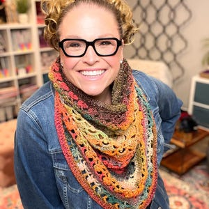 May include: A triangular crochet shawl featuring a variegated colour palette of orange, brown, green, and cream tones. The accessory is crafted with an openwork stitch pattern, creating a textured, lacy appearance that drapes around the neck.