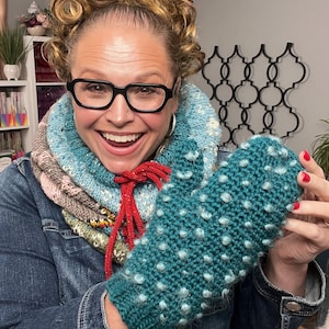 May include: Teal knitted mitten with white bobbles. The mitten is held up by a person wearing a blue and multicolored cowl and a denim jacket. The mitten is a handmade item.